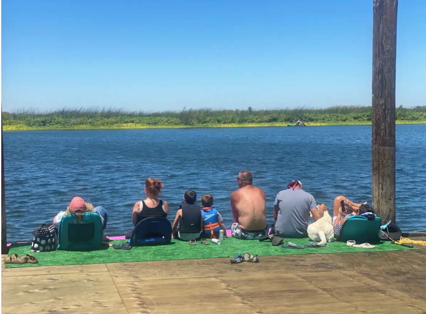 Un groupe de personnes est assis sur un quai, au bord d’un lac paisible. On aperçoit, au premier plan, une zone herbeuse et, à droite, un poteau en bois.