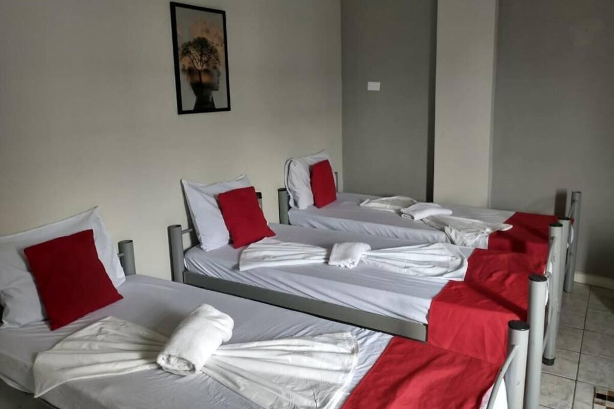 Ein Hotelzimmer mit drei Betten, jedes mit weißen Laken und roten Kissen. Über den Betten hängt ein Gemälde an der Wand.