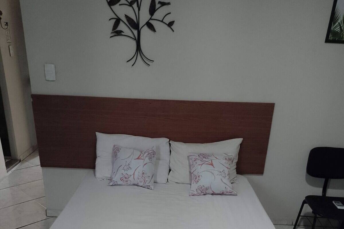 Ein ordentlich gemachtes Bett mit Blumenmuster-Überwurf und roter Decke, ein Holz-Kopfstück und eine Pflanzen-Wanddekoration.