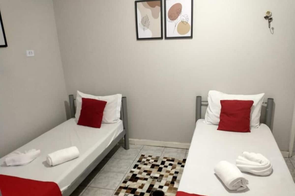 Ein kleines Hotelzimmer mit zwei Einzelbetten, jeweils mit einem roten Kissen und weißer Bettwäsche. Auf den Betten liegen Handtücher und auf dem Boden liegt ein Teppich mit Muster.