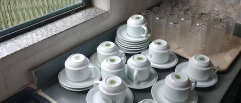 A kitchen counter with stacks of white teacups and saucers, and a row of clear glass tumblers.
