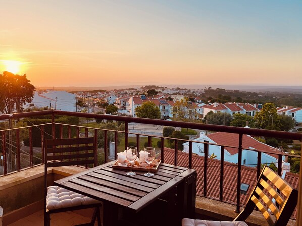A balcony with a table set for two, overlooking a residential area during sunset. - Holiday apartment or home office with a view of Lisbon & the Atlantic coast. (Sesimbra)