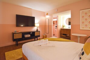 Une chambre d’hôtel avec un lit, une télévision, une table de chevet, un miroir et une salle de bain visible à travers une porte ouverte.
