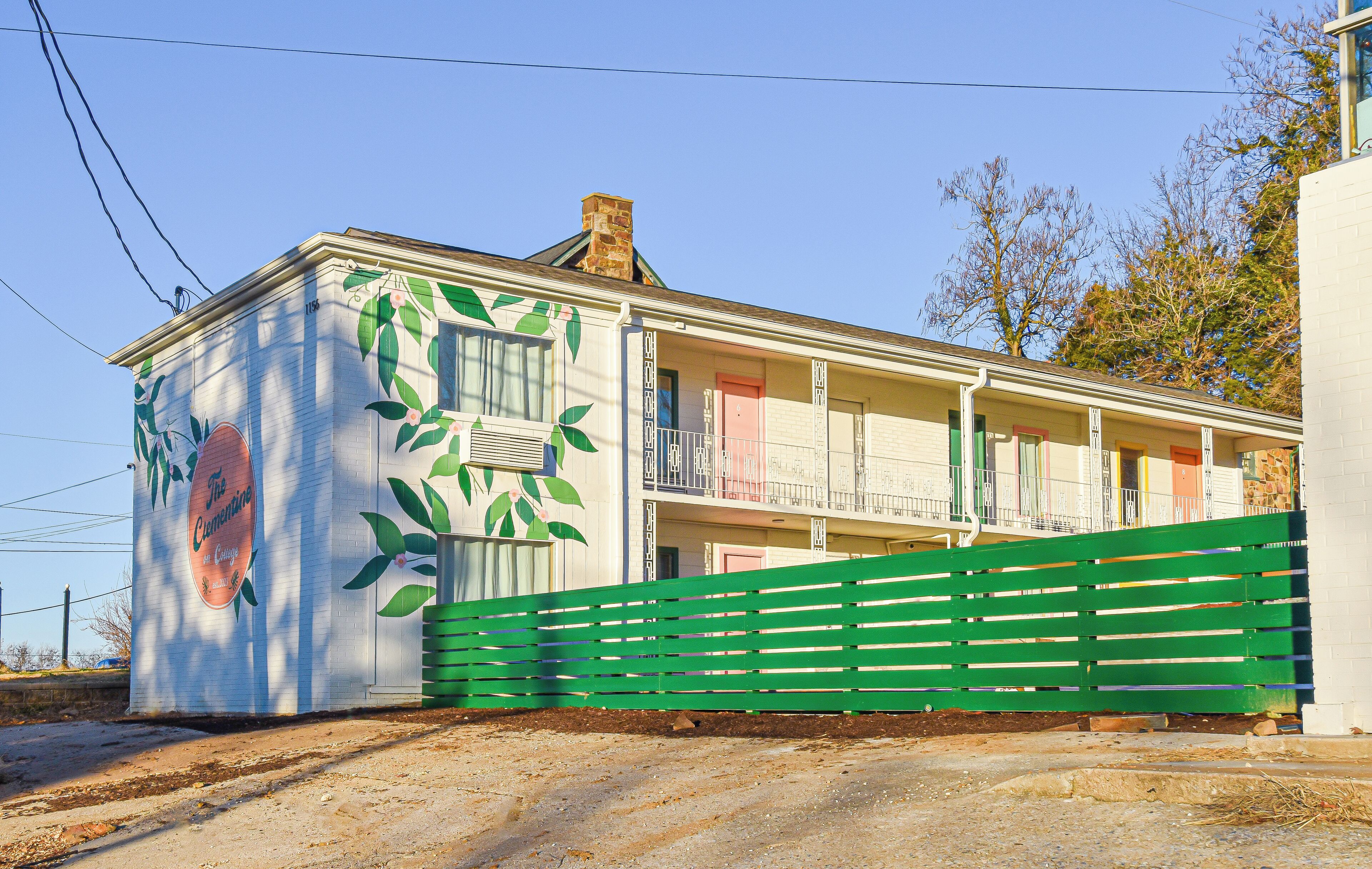 Un edificio con un mural de frutas y hojas en su fachada, una cerca verde en primer plano y un cielo azul despejado al fondo.