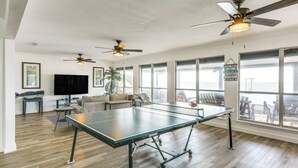 Ein geräumiges Wohnzimmer mit Tischtennisplatte, Deckenventilatoren, großen Fenstern und einem Fernseher.