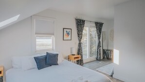 A modern bedroom with a large bed, a small side table, a window with blinds, and a view of the outdoors.