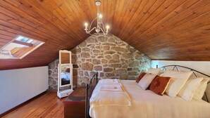 Une chambre avec un mur en pierre, un plafond en bois, un lit avec des oreillers, une table de chevet et un miroir.