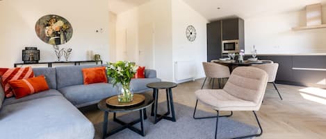 Una sala de estar moderna con un sofá gris, cojines rojos, una mesa de centro y una silla. Se ve un área de comedor con una mesa y sillas al fondo.