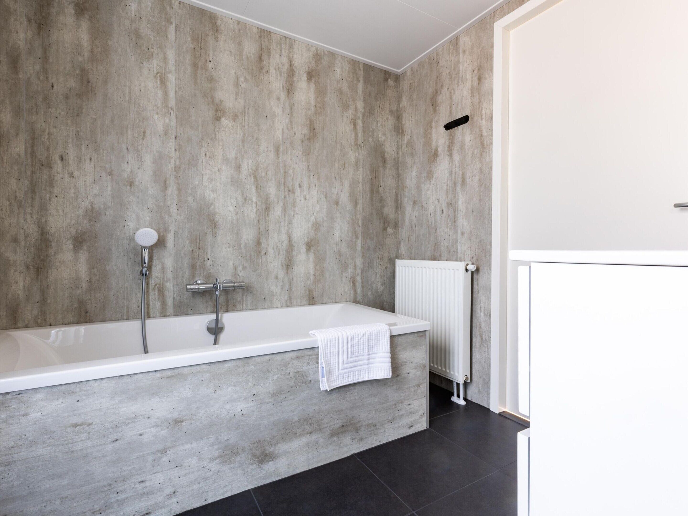 A modern bathroom with a concrete wall, a white bathtub, and a white radiator.