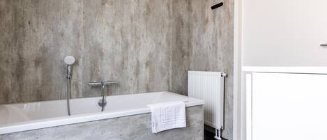 A modern bathroom with a concrete wall, a white bathtub, and a white radiator.