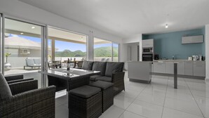 Uma cozinha moderna com armários brancos e uma parede azul de destaque, uma área de jantar com mobiliário de vime e uma mesa de vidro, e vista para colinas verdes através de grandes janelas.