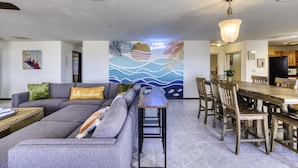A modern living room with a grey sectional sofa, a wooden dining table with chairs, and a wall mural with a tropical theme. - 99 Problems & the Beach Ain`t 1 (Daytona Beach)