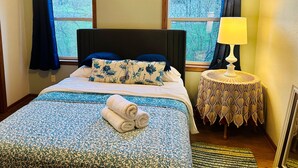 Una cama bien tendida con una colcha y almohadas de estampado floral, una mesita de noche con mantel de encaje y una ventana con cortinas.