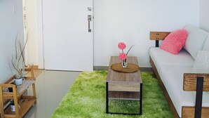 Uma sala de estar moderna com um sofá, uma mesa de centro com um vaso de flores, um tapete verde e uma televisão na parede.