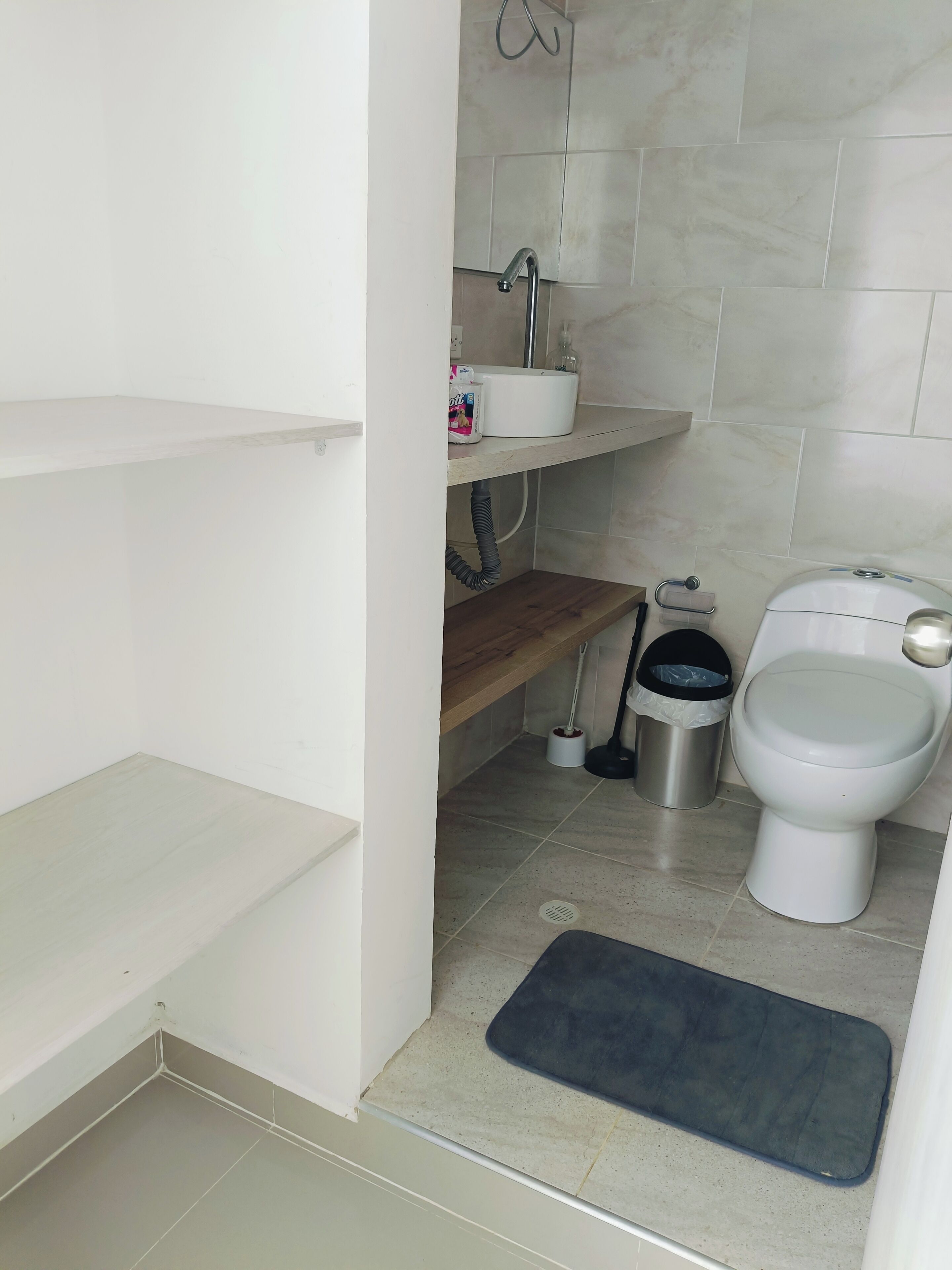 A bathroom with a toilet, a sink, a shelf with toiletries, a trash bin, and a mat on the floor.