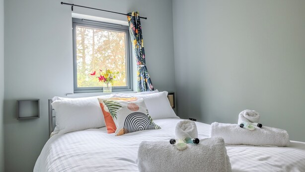 Ein ordentlich bezogenes Bett mit weißer Bettwäsche und Zierkissen, ein Fenster mit Blick auf Bäume sowie ein Handtuch mit einer kleinen Dekoration.