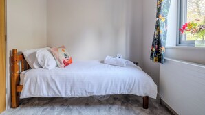Ein kleines, ordentlich gemachtes Bett mit weißer Bettwäsche, einigen Kissen, einem hölzernen Kopfteil und einem Fenster mit floralem Vorhang.