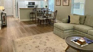 Ein modernes Wohnzimmer mit einer Eckcouch, einem Essbereich mit Tisch und Stühlen, einem Deckenventilator und einer Küche im Hintergrund.
