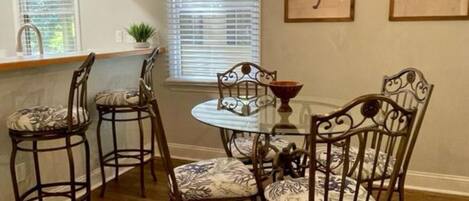 A dining area with a round table and four chairs, wooden flooring, pendant lights, a window with blinds, and two framed artworks on the wall.