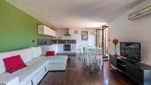 A modern living room with a white sofa, red cushions, a dining area with white chairs, a black entertainment unit with a TV, and a kitchen in the background.