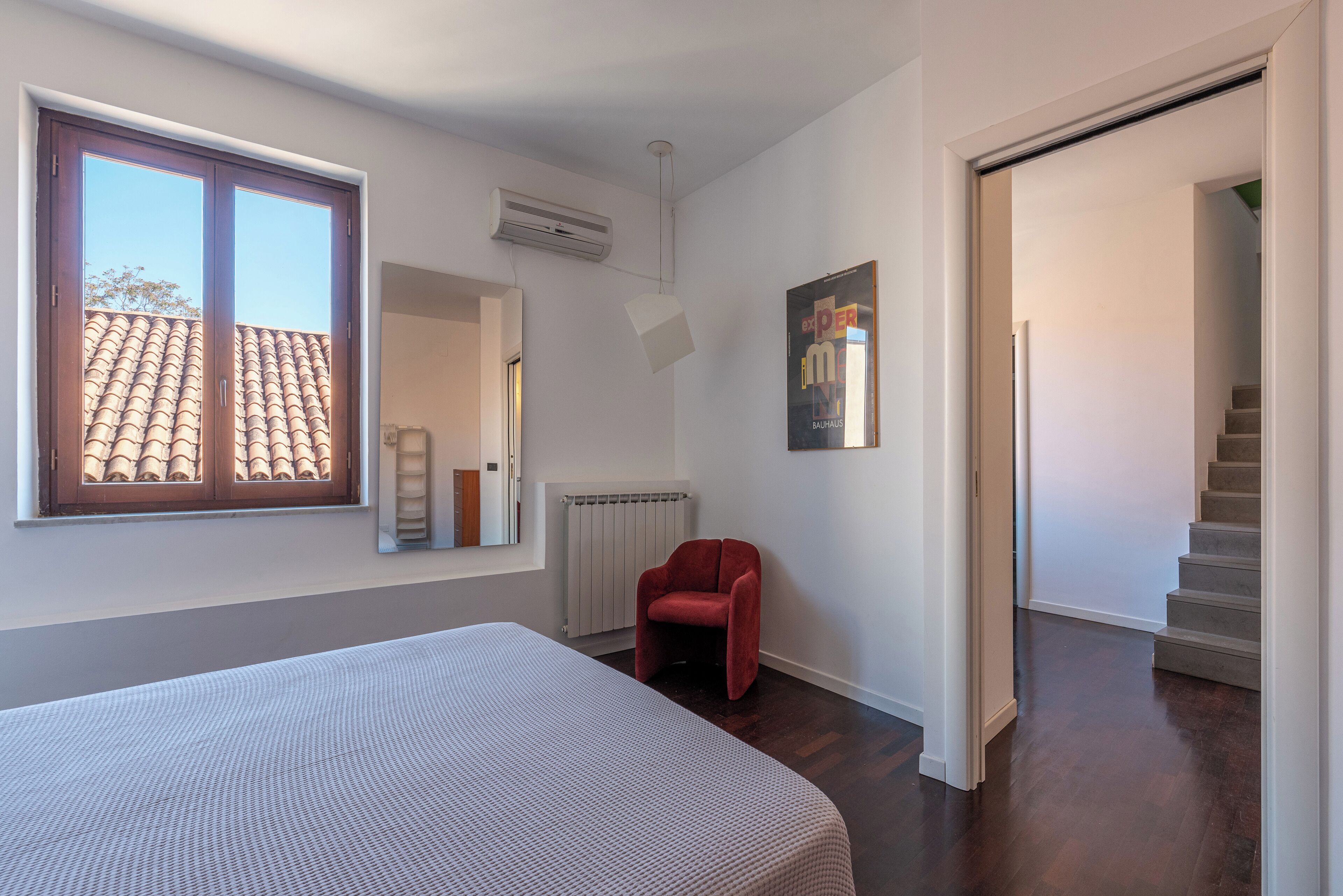 Un dormitorio con una cama grande, una silla roja, una ventana con vista a un tejado de tejas, aire acondicionado y un póster en la pared.