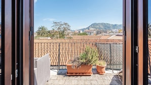 A balcony with terracotta tiles, potted plants, and a view of a cityscape with mountains in the distance.