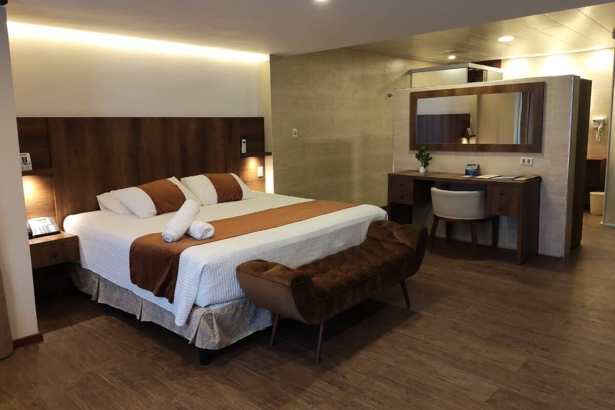 A hotel room with a large bed, a desk, a chair, a vanity with a mirror, and a bench.