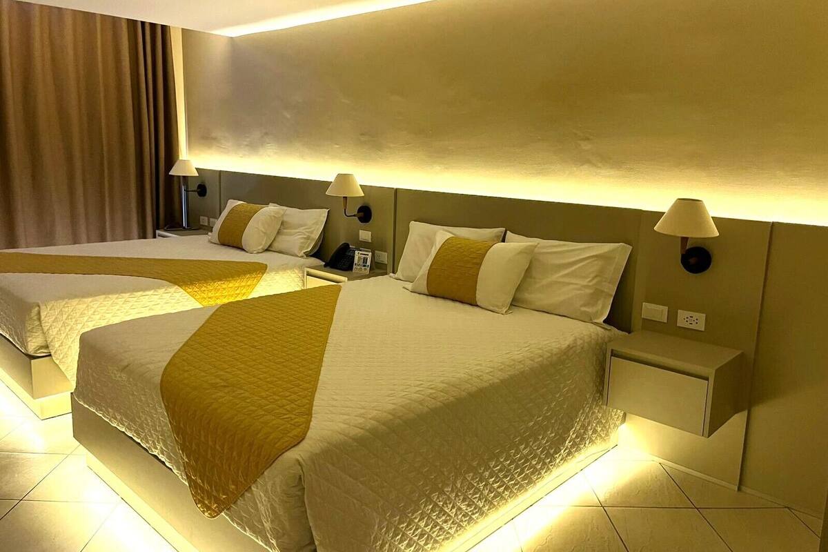 A hotel room with two beds, each with a headboard, bedside tables, lamps, and a curtain.