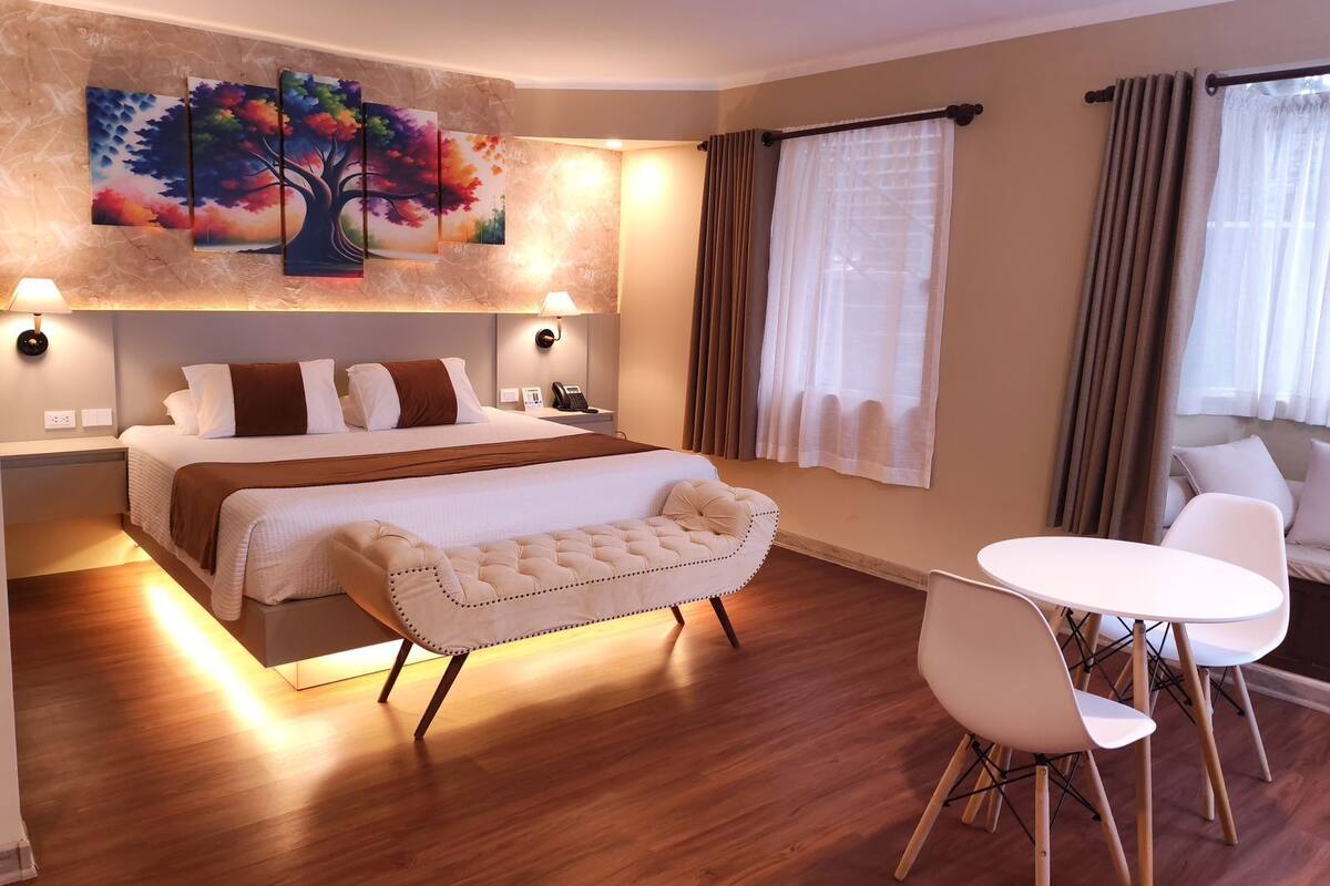 A modern hotel room with a large bed, a sofa, a dining table with chairs, and a painting of a tree on the wall.