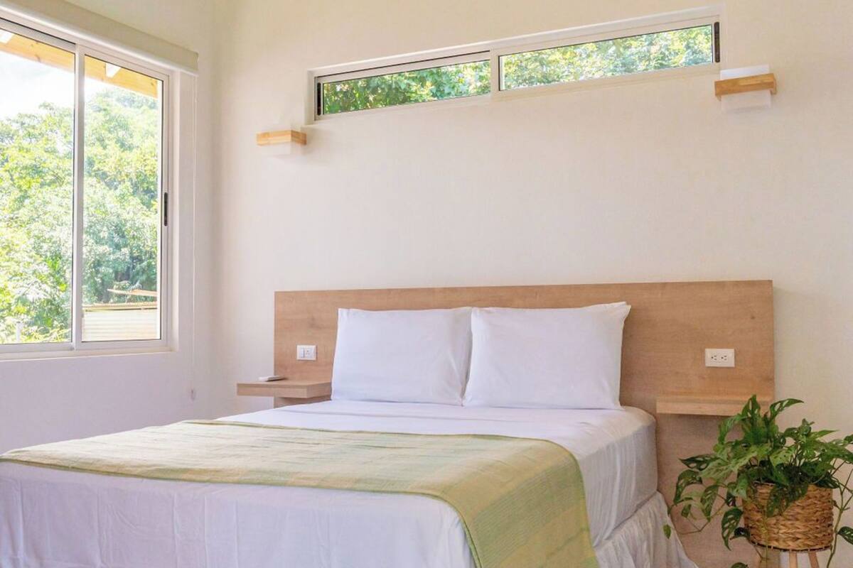 A bedroom with a wooden ceiling, a large window, a bed with white bedding, a bedside table, and a potted plant.
