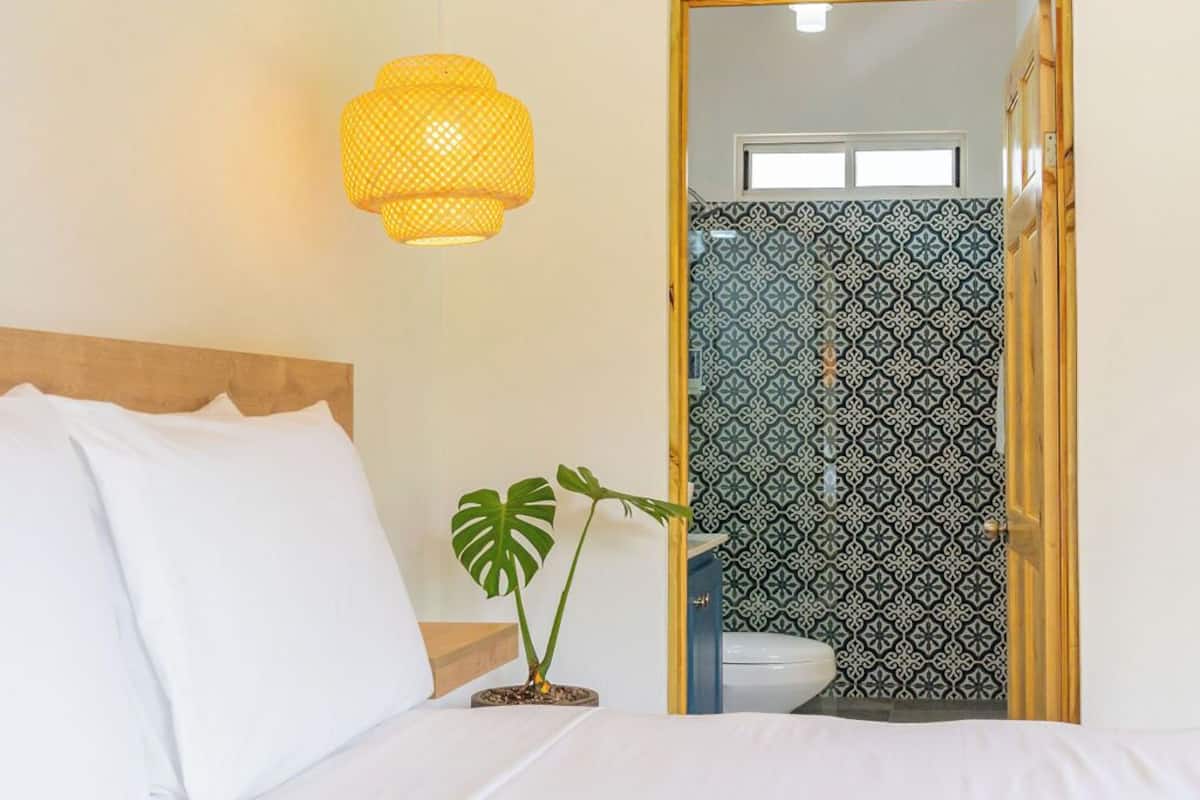 A bedroom with a bed, a hanging light fixture, a potted plant, and a bathroom visible through an open door.