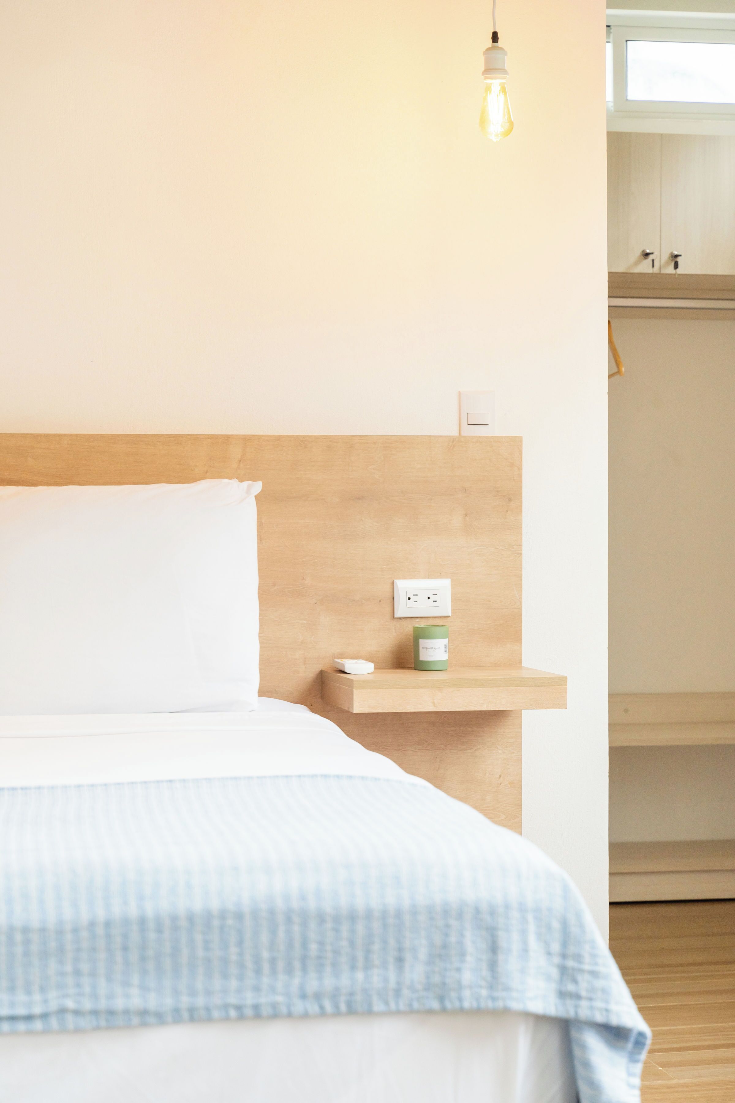 A neatly made bed with a white pillow and a blue checkered blanket, a bedside table with a green container, and a light bulb hanging above.