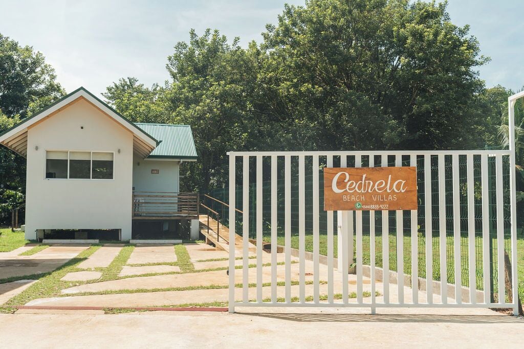 Uma entrada protegida por portão para um imóvel chamado "Cedrela Beach Villas", com cerca branca e uma placa.
