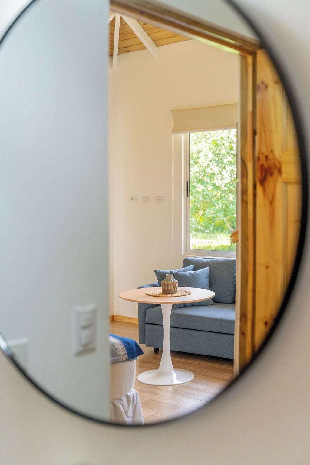 Ein Zimmer mit einem runden Spiegel, der ein blaues Sofa, einen kleinen runden Tisch und ein Fenster mit Blick auf Grünflächen reflektiert.