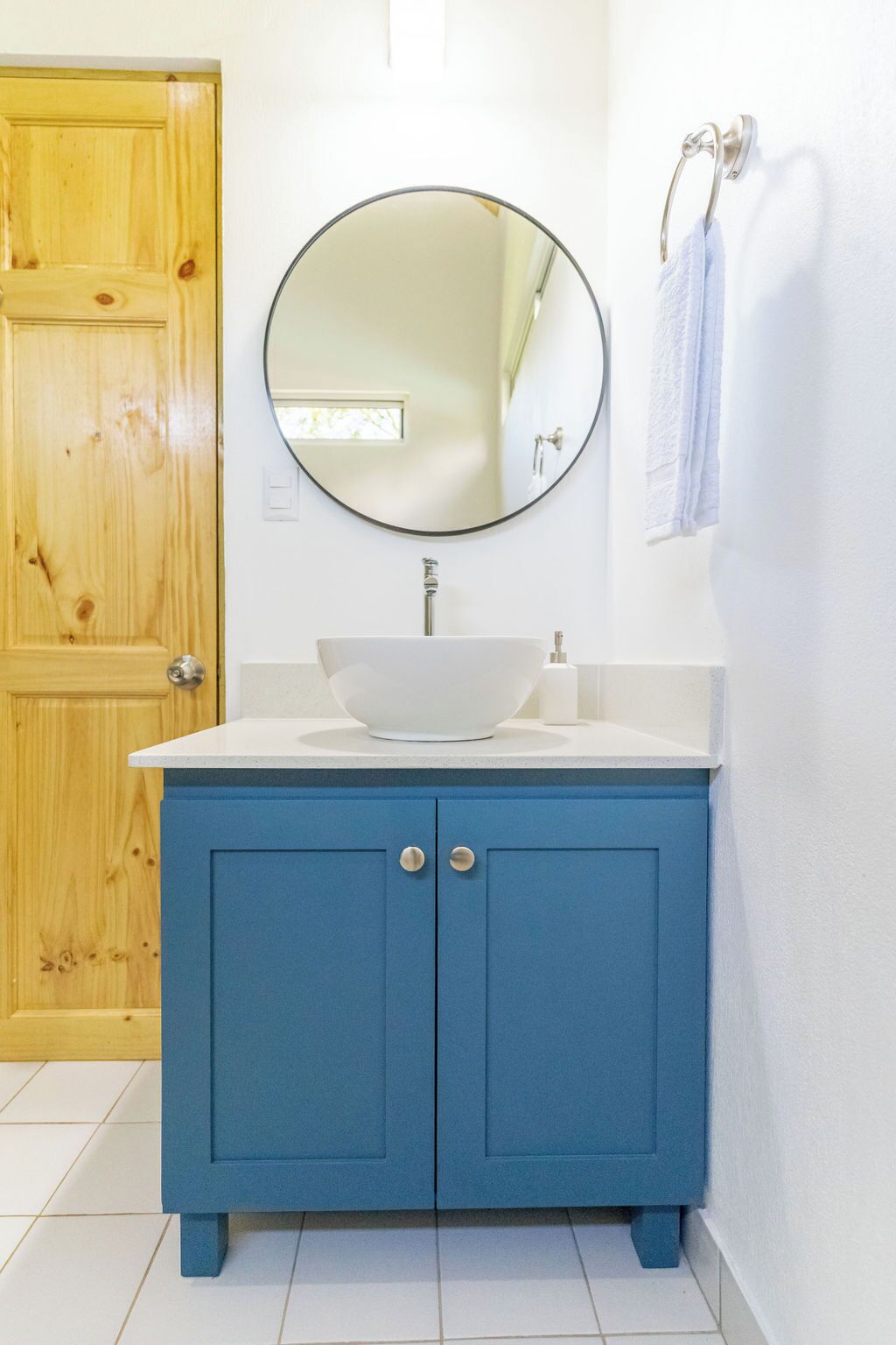 A bathroom with a wooden door, a white sink with a round mirror above it, and a blue cabinet.