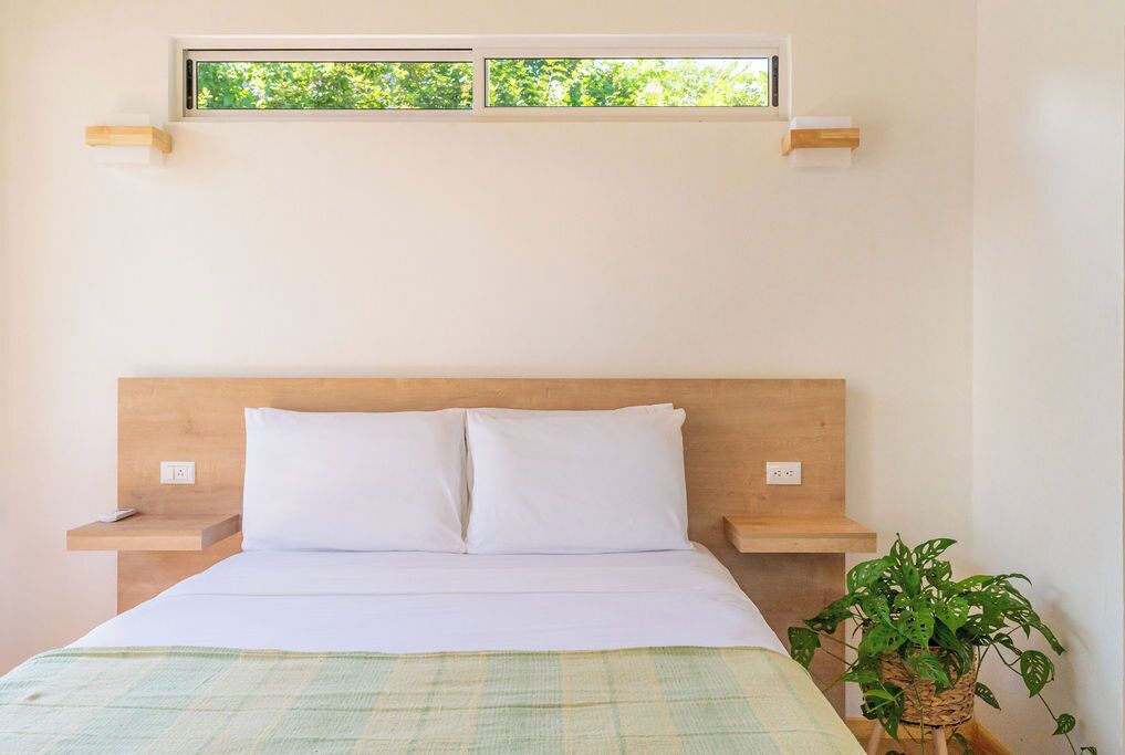Een bed met witte lakens, een houten hoofdbord, nachtkastjes en een raam met uitzicht op groen.