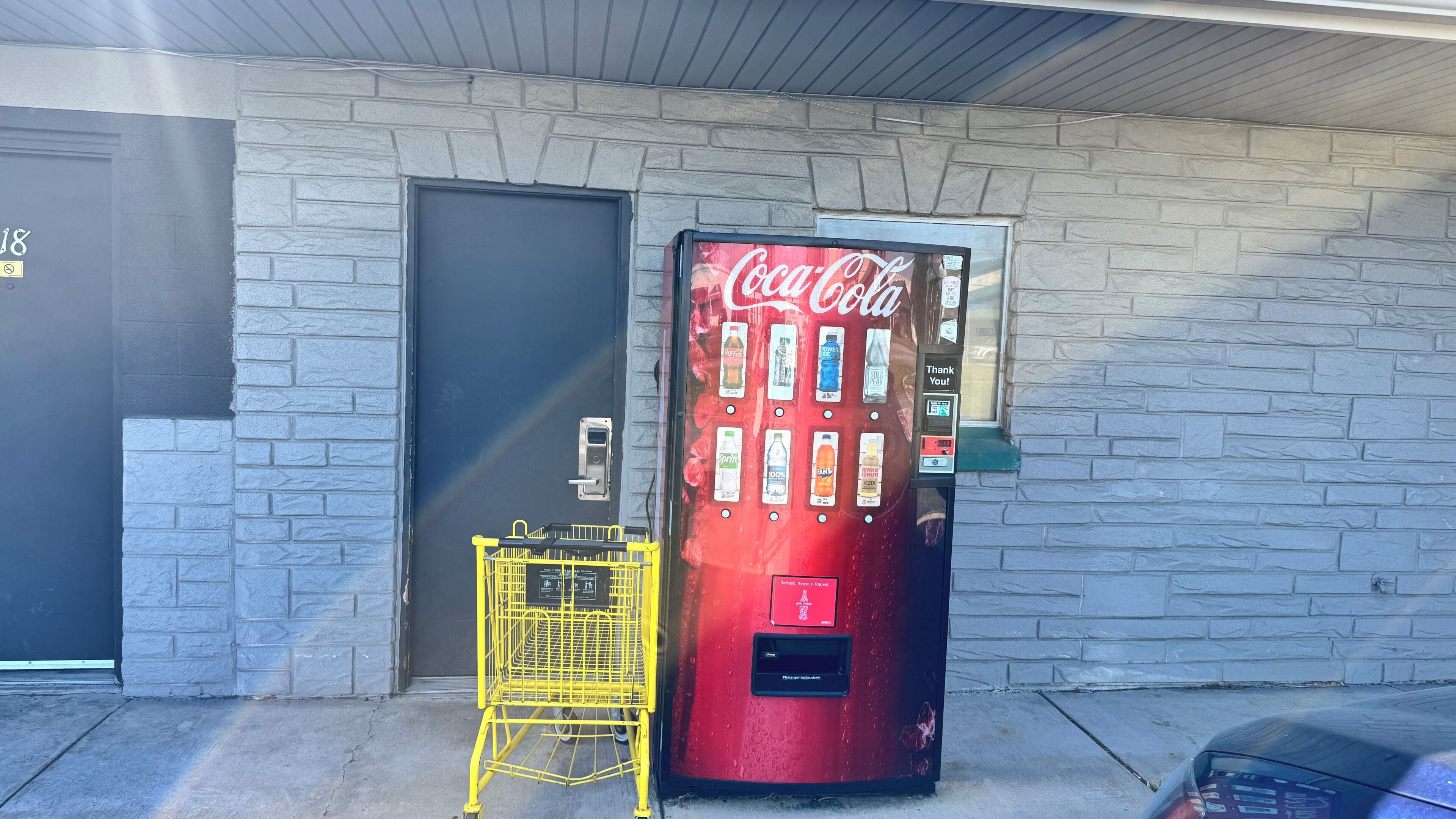 Een Coca-Cola automaat naast een deur met een gele winkelwagen.