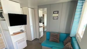 A compact living space with a teal sofa, a flat-screen TV mounted on a white cabinet, and a kitchen area visible through an open door. - Mobile home sea view high up on the Opal Coast  (Le Portel)