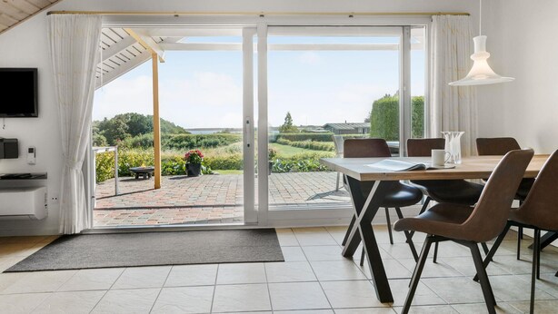 A dining area with a table and chairs, a TV mounted on the wall, and a view of a garden and buildings through large glass doors. - 3 bedroom nice home in Egernsund (Egernsund)