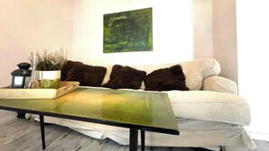 Una sala de estar con un sofá blanco, una mesa de centro de vidrio, un cuadro abstracto verde y una planta pequeña en maceta.