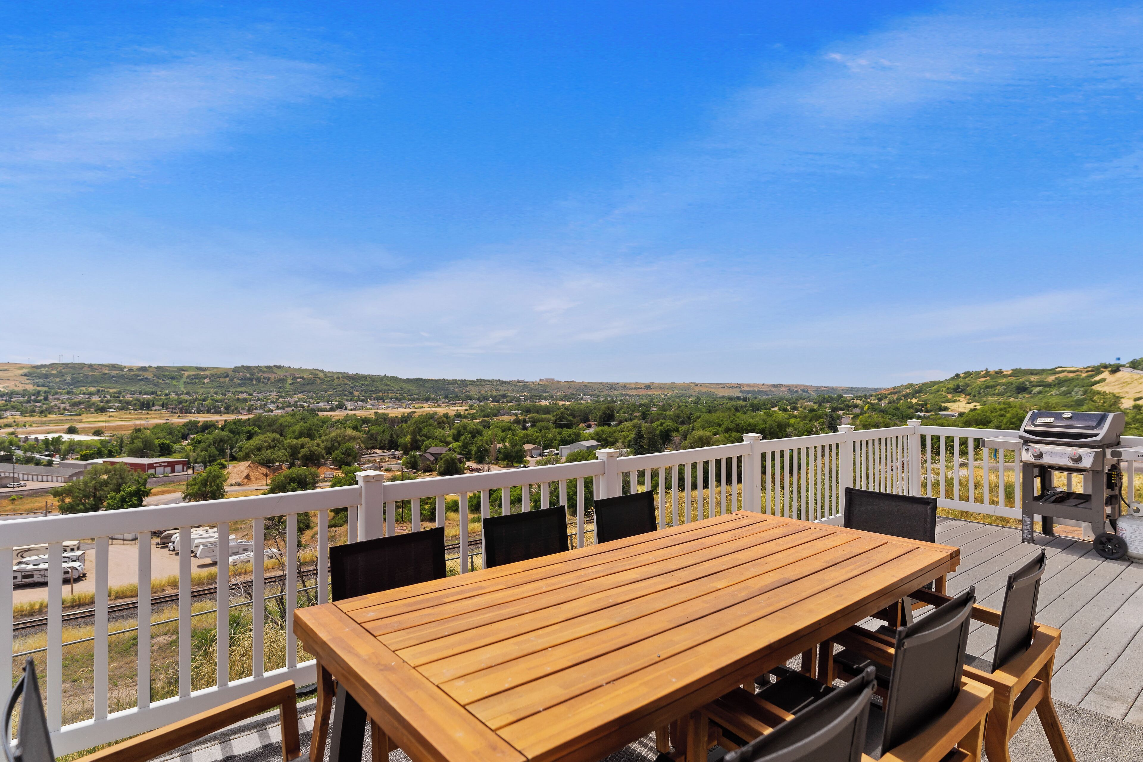 An outdoor deck with a wooden table and chairs, a barbecue grill, and a white fence overlooking a scenic landscape.