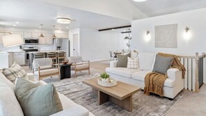 A modern living room with a sofa, coffee table, and chairs. There is a kitchen in the background with cabinets and appliances.