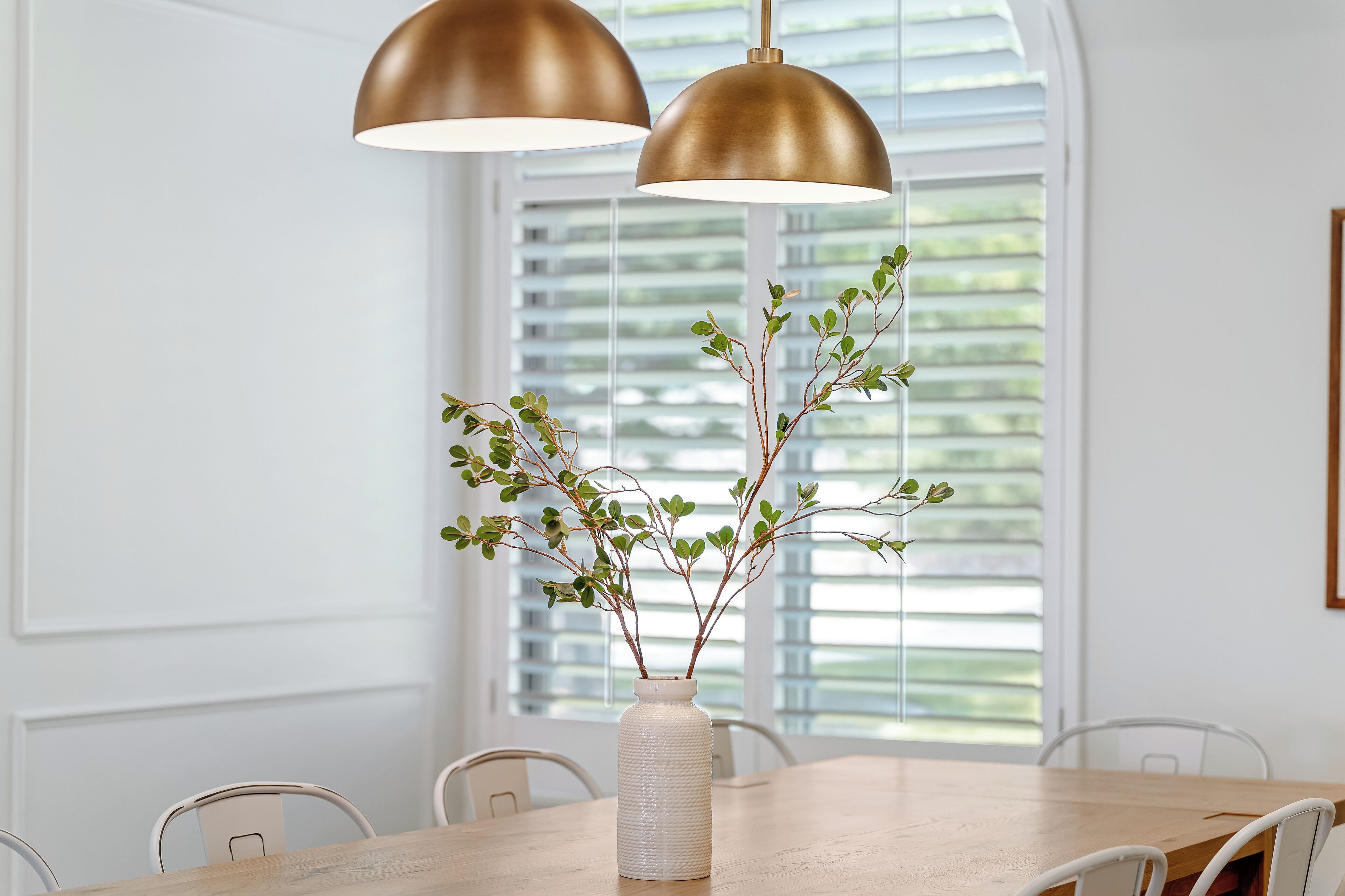 A dining area with a wooden table, white chairs, a white vase with green branches, and two gold pendant lights.