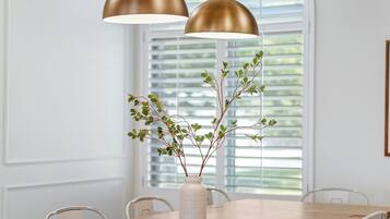 A dining area with a wooden table, white chairs, a white vase with green branches, and two gold pendant lights.