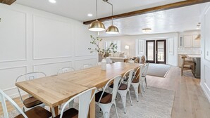 A spacious dining area with a wooden table and white chairs, a kitchen in the background, and pendant lighting.