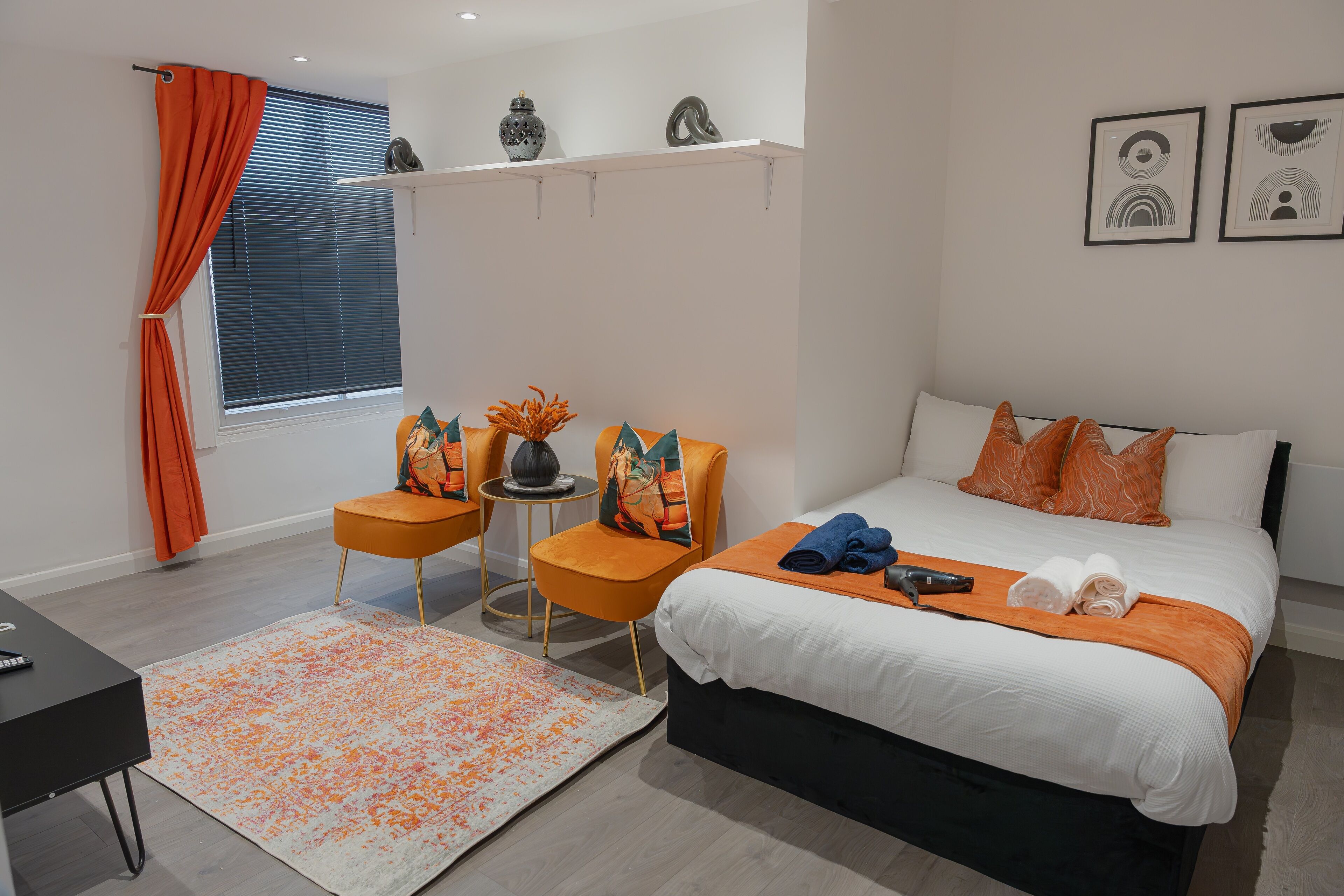 A modern bedroom with a large bed, orange accent pillows, a side table with a vase, and a view of the outdoors through a window with blinds.