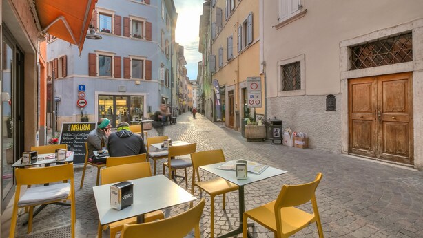 A café with outdoor seating, tables, and yellow chairs, set against a cobblestone street with buildings on either side. - Casa Arx in Arco With 1 Bedrooms and 1 Bathrooms (Arco)