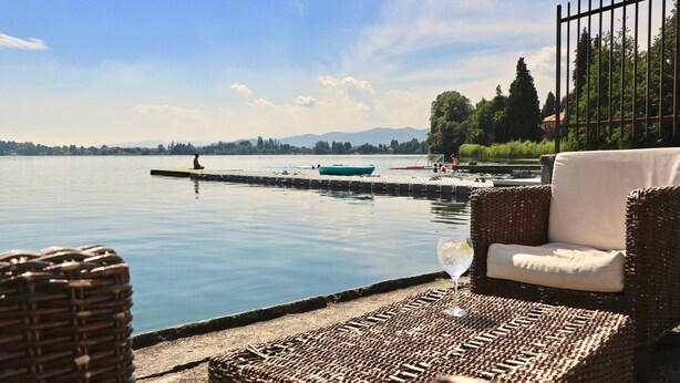 A serene lakeside setting with a wicker lounge chair and a glass of wine, overlooking a calm lake with a distant mountain range and a small boat on the water. - Riva del Caolino in Ispra (Ispra)