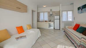 Un espacio habitable compacto con kitchenette, un sofá con cojines de colores y una cama con cubrecama blanco.