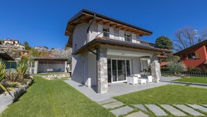Ein zweistöckiges Haus mit Balkon, einer Terrasse und Gartenmöbeln sowie einer Steinpfeiler.